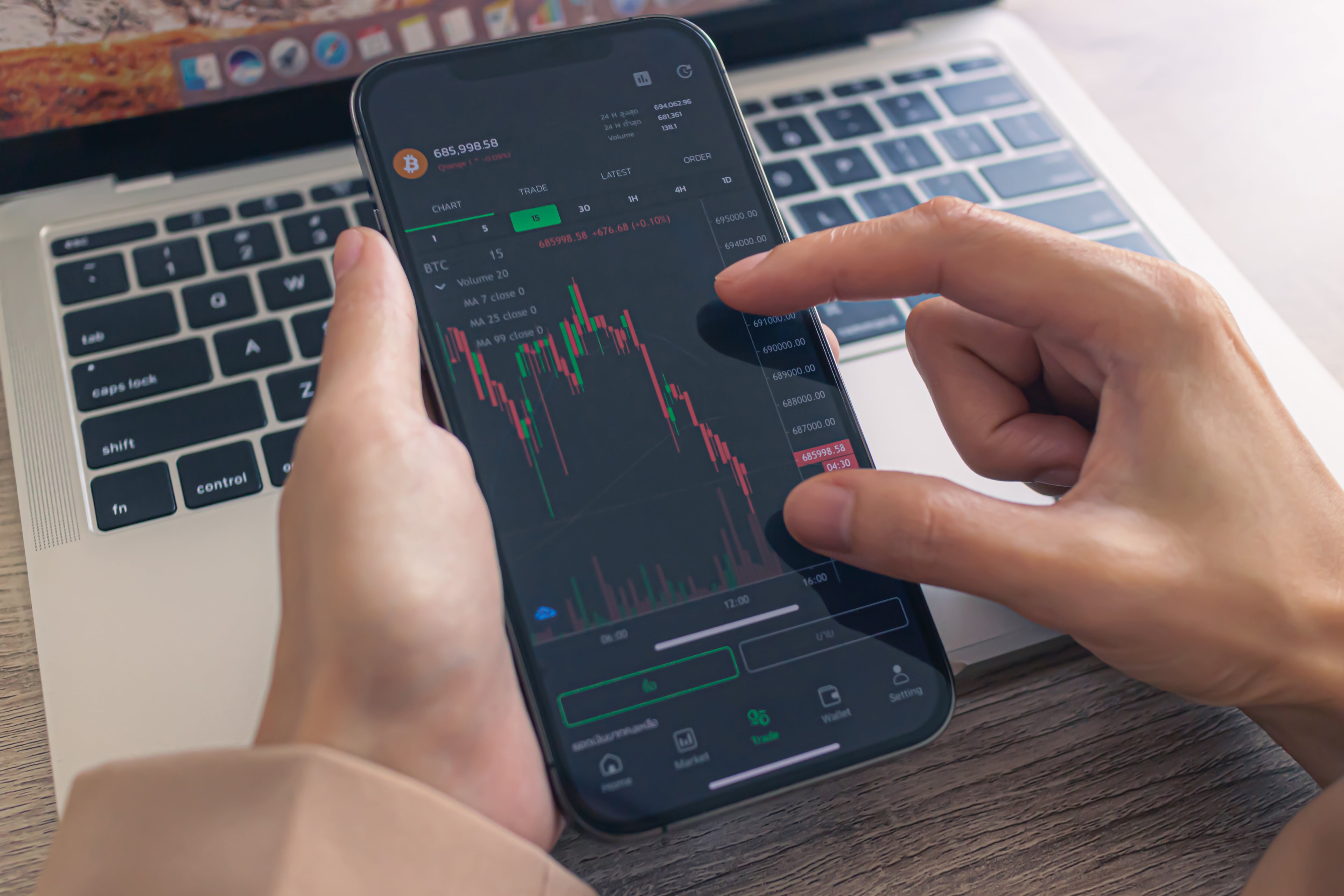 A woman trading bitcoin on her mobile device infront of a laptop from her POV.